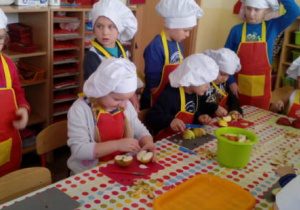 Grupa kucharzy przy stole kroi jabłka, pozostali obserwują technikę krojenia.